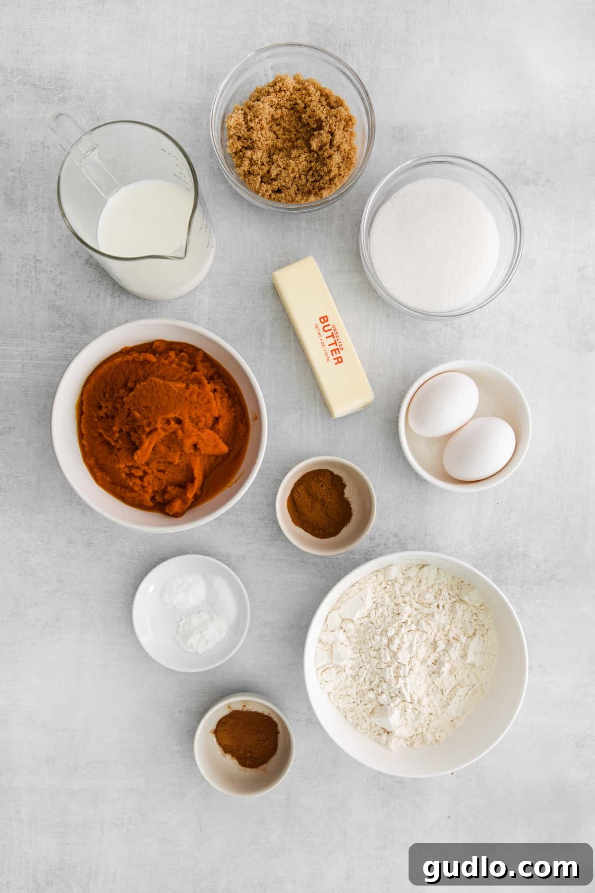 A vibrant collection of fresh ingredients for both pumpkin bars and their cream cheese frosting, meticulously arranged in bowls on a white surface, ready for the baking process.