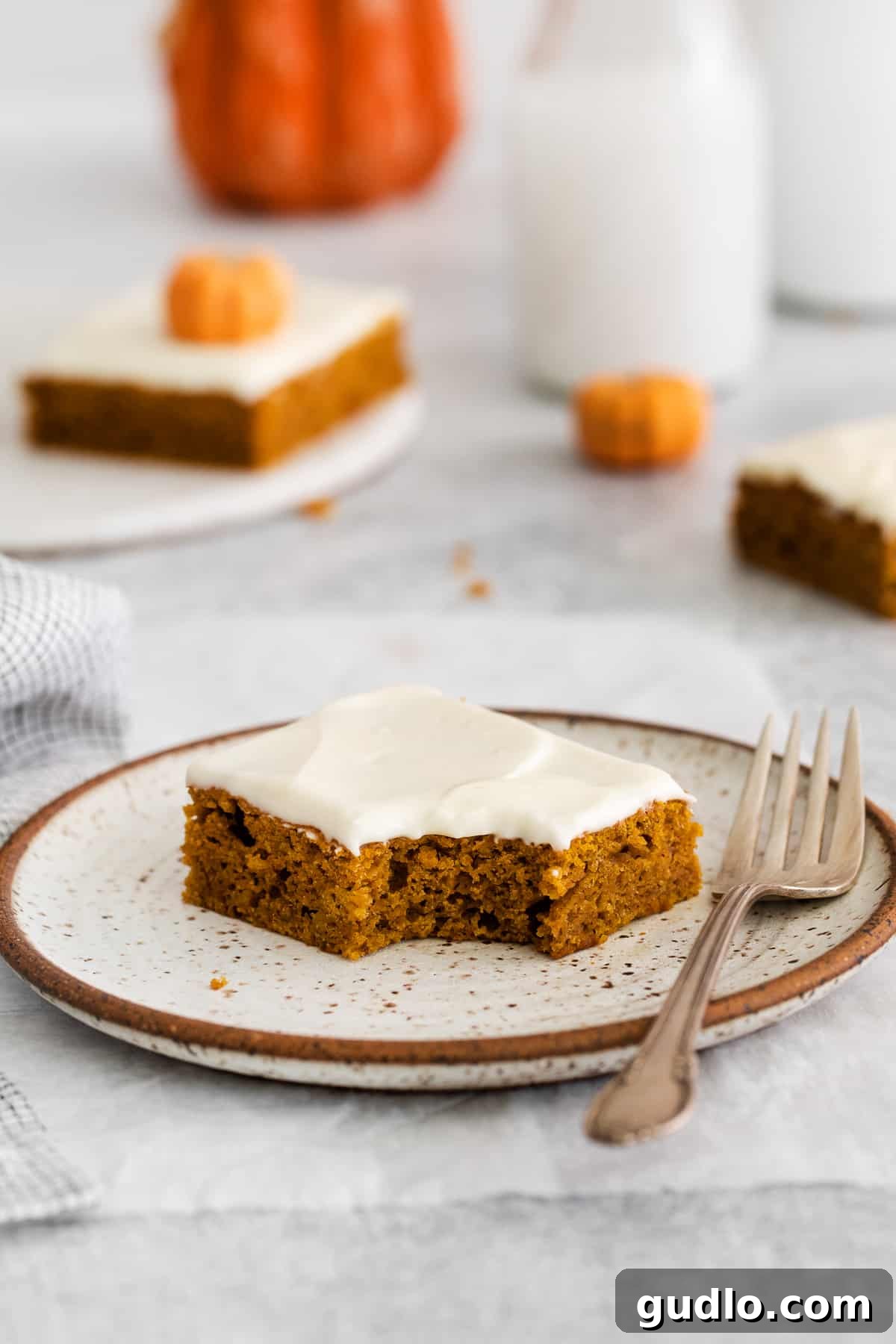 A single pumpkin bar on a plate with a noticeable bite taken out, revealing its moist interior and the thick, creamy layer of frosting.