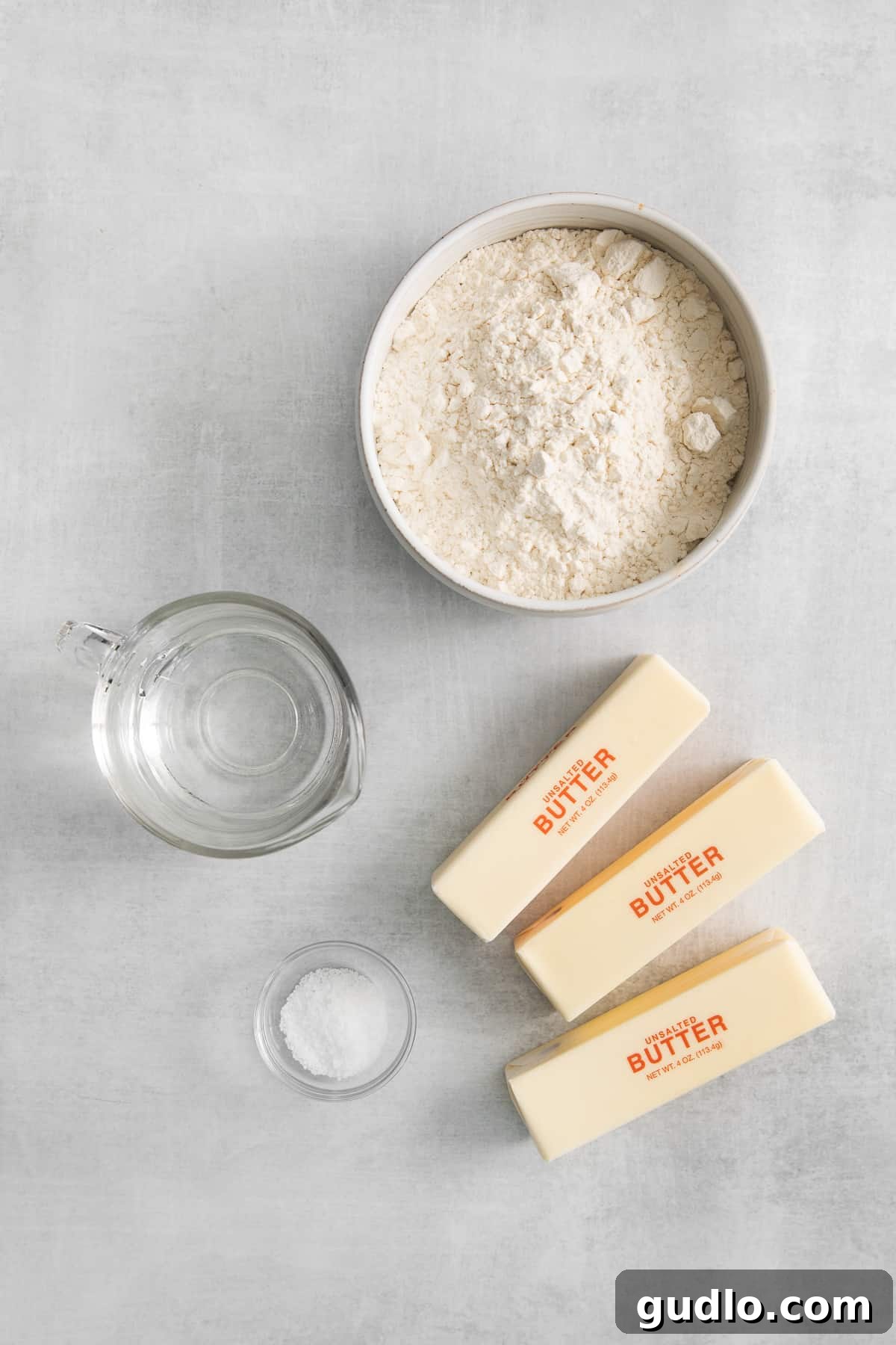 The four essential ingredients for crafting homemade puff pastry: all-purpose flour, a sprinkle of kosher salt, generous blocks of cold unsalted butter, and a glass of ice water, neatly arranged on a kitchen counter.