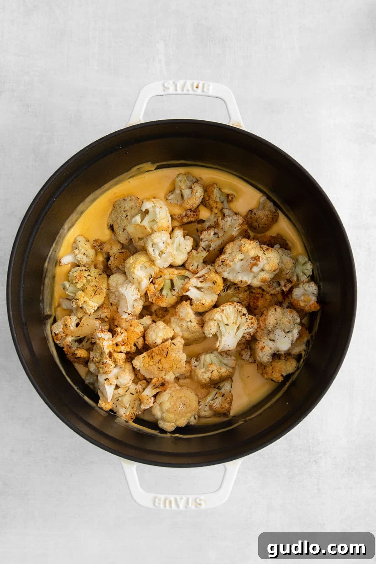 Roasted cauliflower florets being poured over the prepared cheese sauce in a large bowl, ready for mixing.