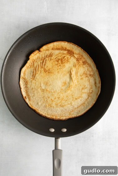A golden pancake cooking in a frying pan on a white background, demonstrating the crepe-making process.