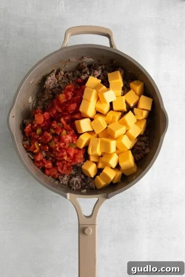 A frying pan with melting Velveeta cheese, ground meat, and tomatoes, actively being stirred.