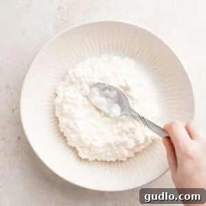 Homestyle Cottage Lasagna 6 A person pouring a spoonful of fluffy white cottage cheese into a small bowl, ready for a recipe.