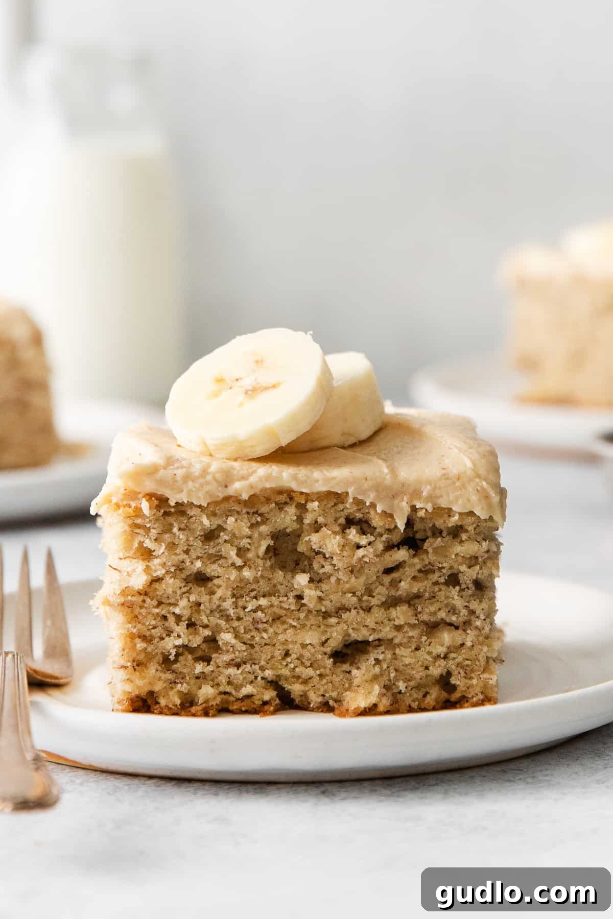 A perfectly sliced piece of moist banana cake, topped generously with creamy peanut butter cream cheese frosting, resting on an elegant white plate, ready to be savored.