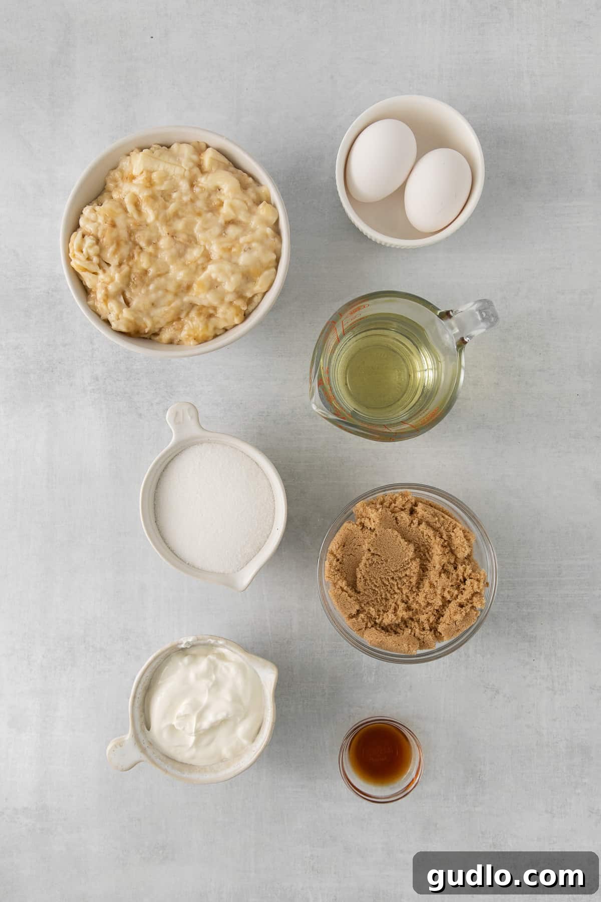 Various banana cake ingredients, including mashed bananas, eggs, flour, and sugar, neatly arranged in separate bowls on a kitchen counter, ready for baking.