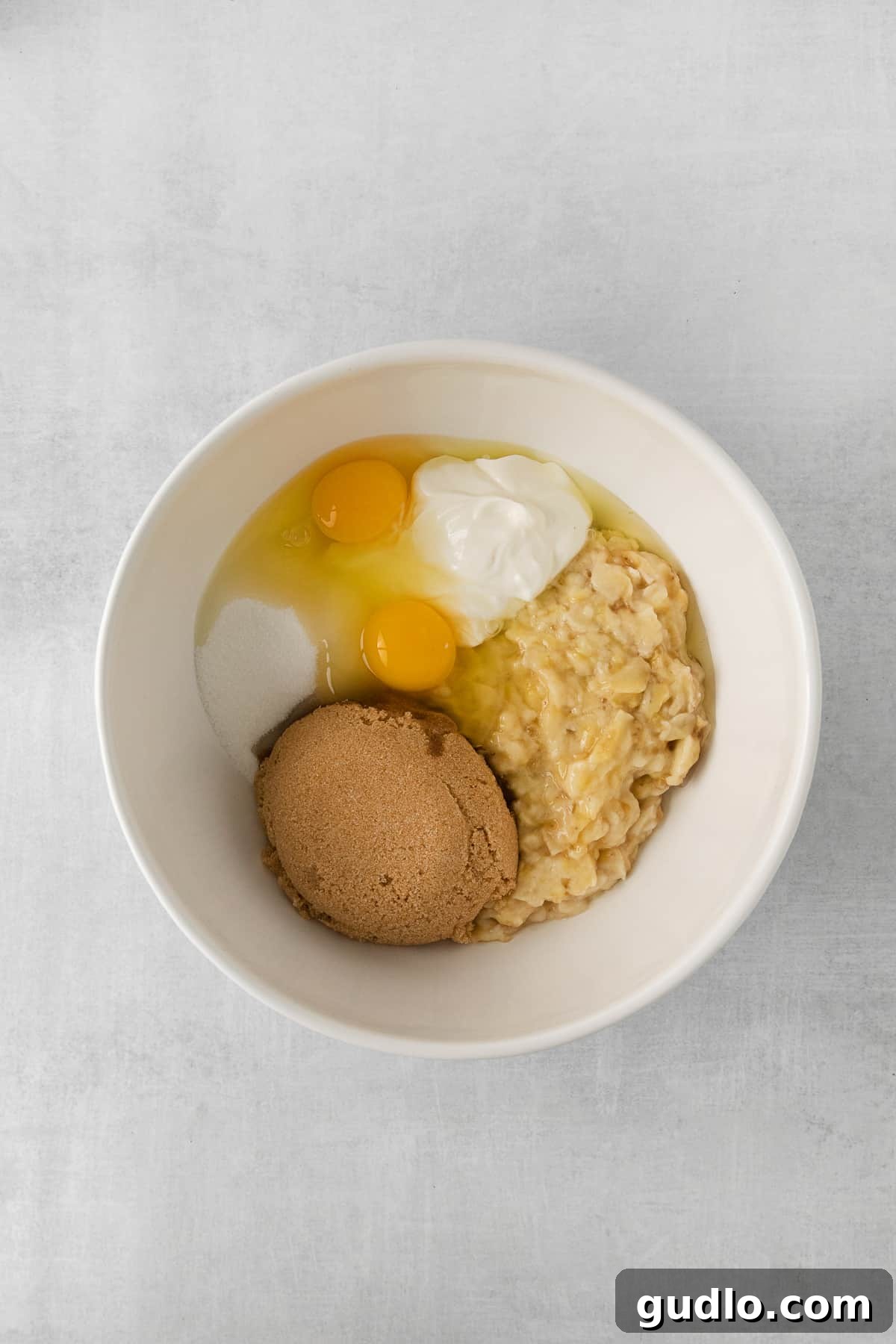 Mashed ripe bananas, along with other wet ingredients like oil, eggs, and sugar, mixed together in a large white bowl, forming the base of the banana cake batter.