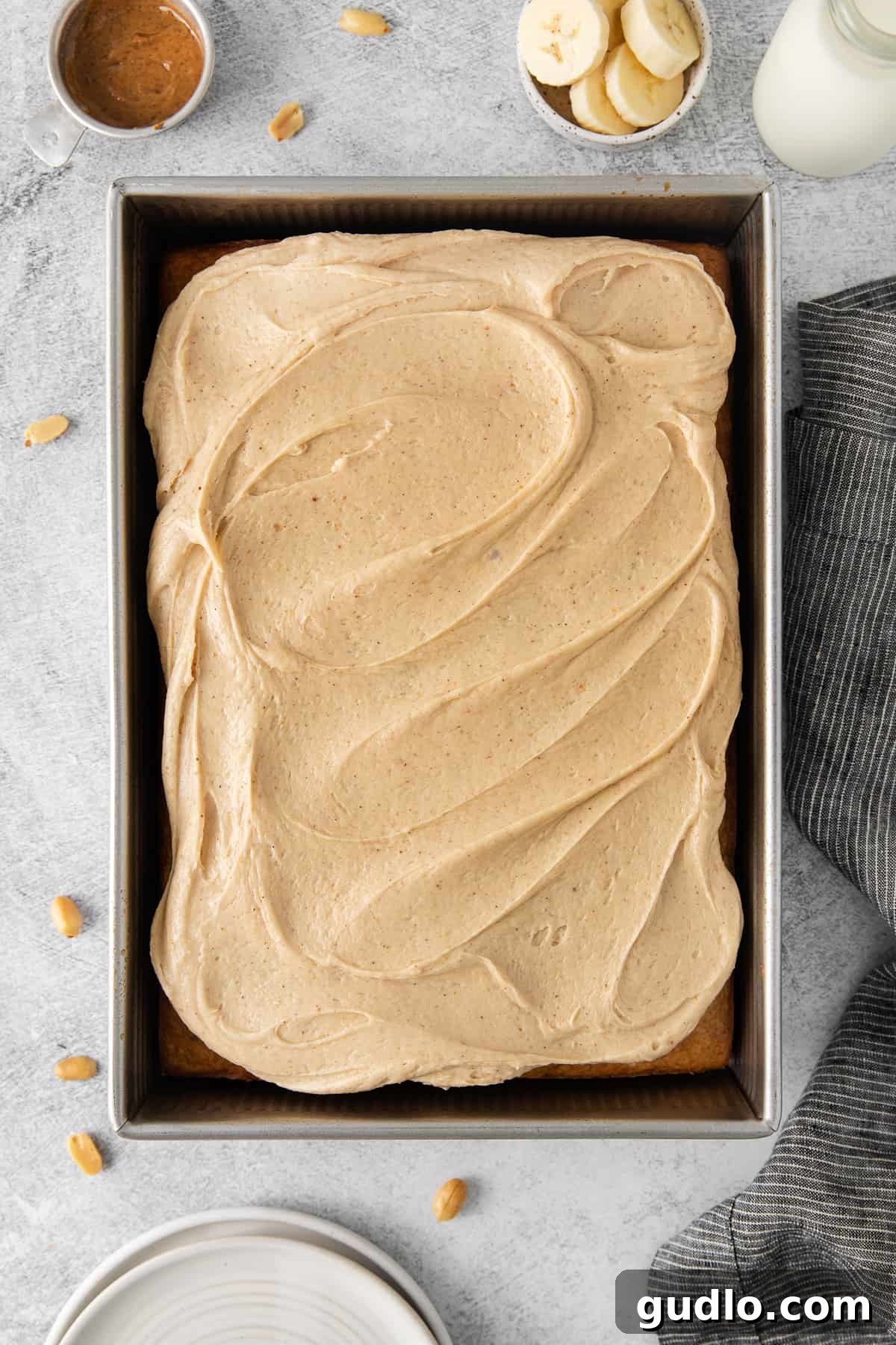 A freshly baked banana cake generously topped with creamy peanut butter cream cheese frosting, still in its baking pan, with a perfectly cut slice ready to be lifted.