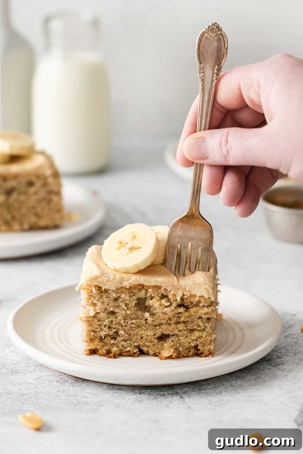A close-up of a single slice of banana cake with a thick layer of creamy peanut butter cream cheese frosting, showing the moist crumb and a hint of banana, on a white plate.