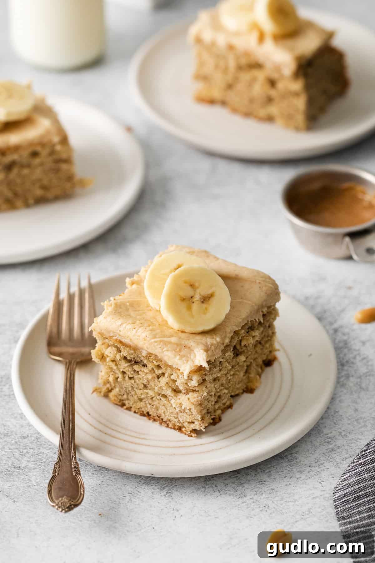 A delicious slice of banana cake, richly covered with peanut butter cream cheese frosting, garnished with a sprinkle of peanuts, served on a white plate with a fork.