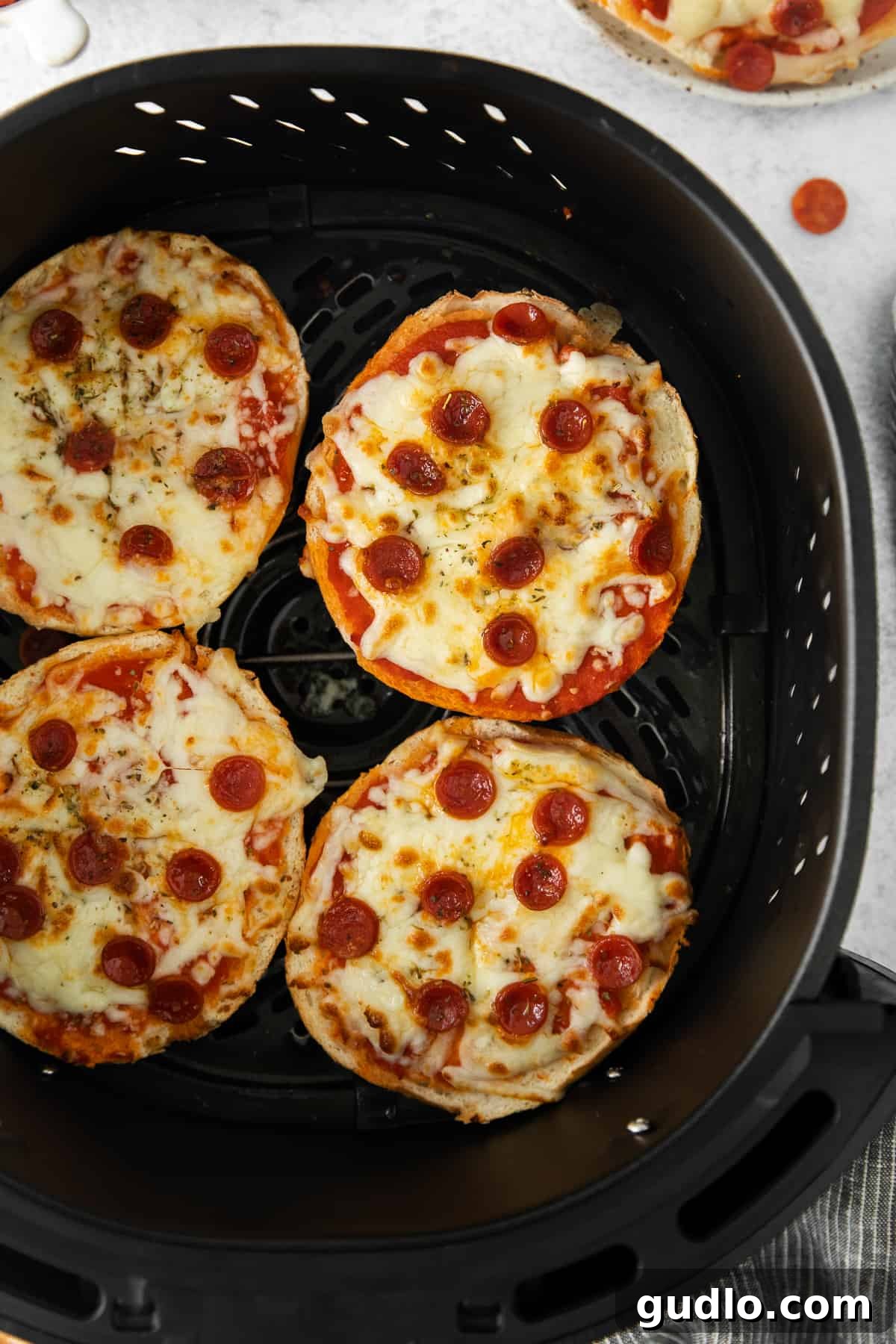 Pizza bagels getting perfectly crispy and cheesy in the air fryer. Two cheesy air fryer pizza bagels, topped with mini pepperoni, cooking to perfection in an air fryer basket, with the cheese bubbling and edges crisping up.