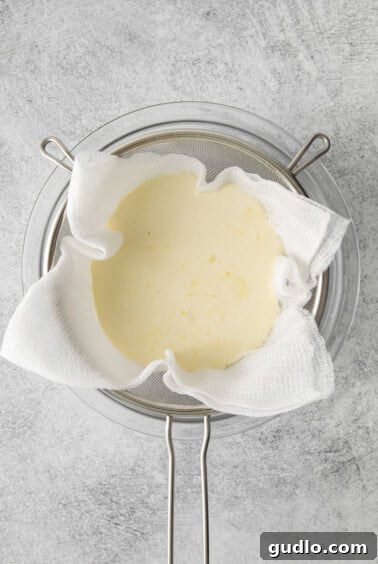 Mascarpone cheese actively draining in a cheesecloth-lined colander within a bowl.
