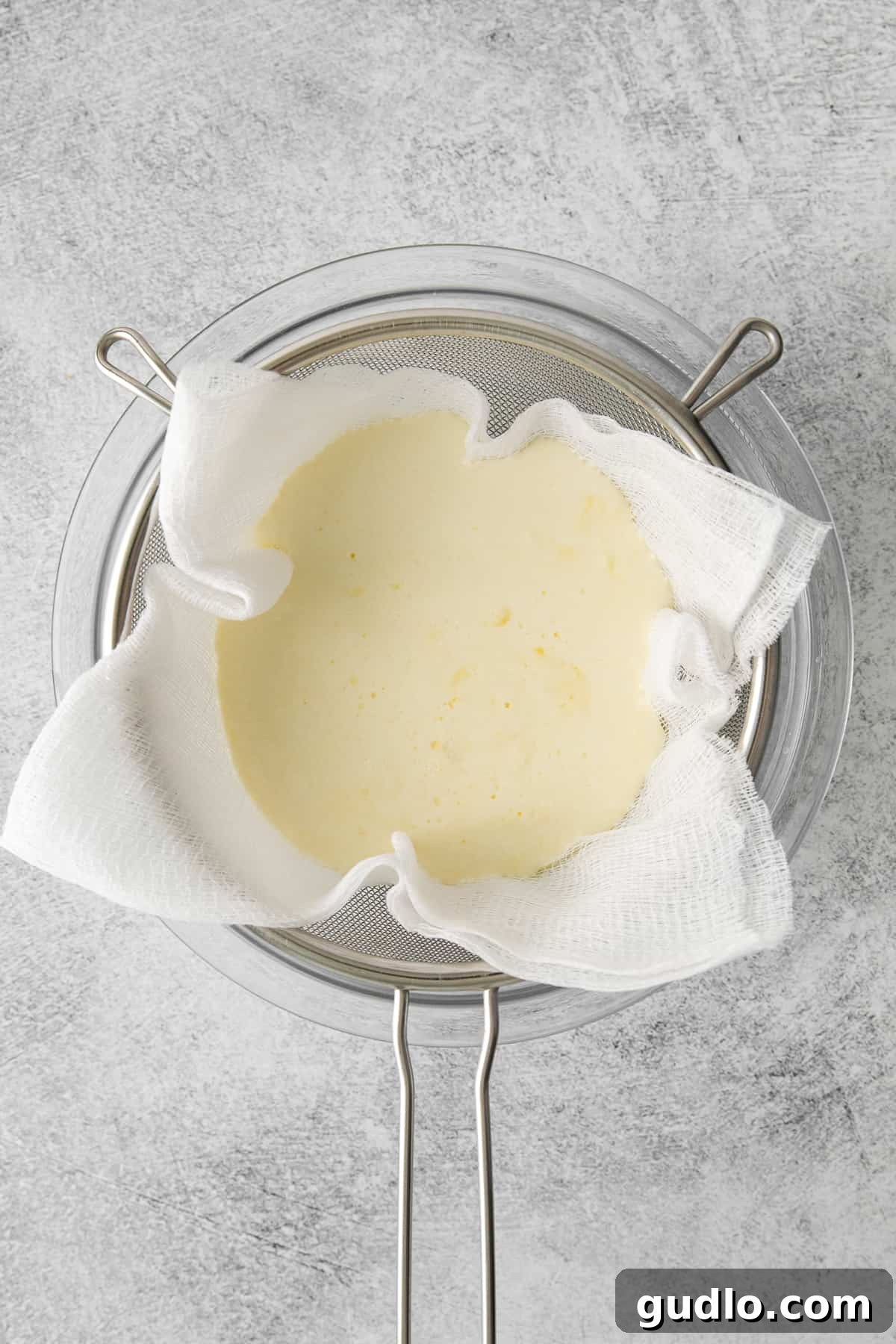 Homemade mascarpone cheese wrapped in cheesecloth, actively draining in a colander over a bowl in the refrigerator.