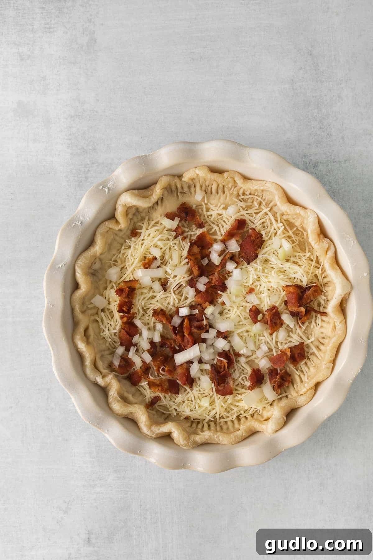 Our easy Quiche Lorraine recipe is the perfect dish for your next brunch! Made with fluffy eggs, cheese, bacon and onions. A parbaked pie crust filled with crumbled bacon, shredded Gruyère cheese, and minced onion, awaiting the egg custard.