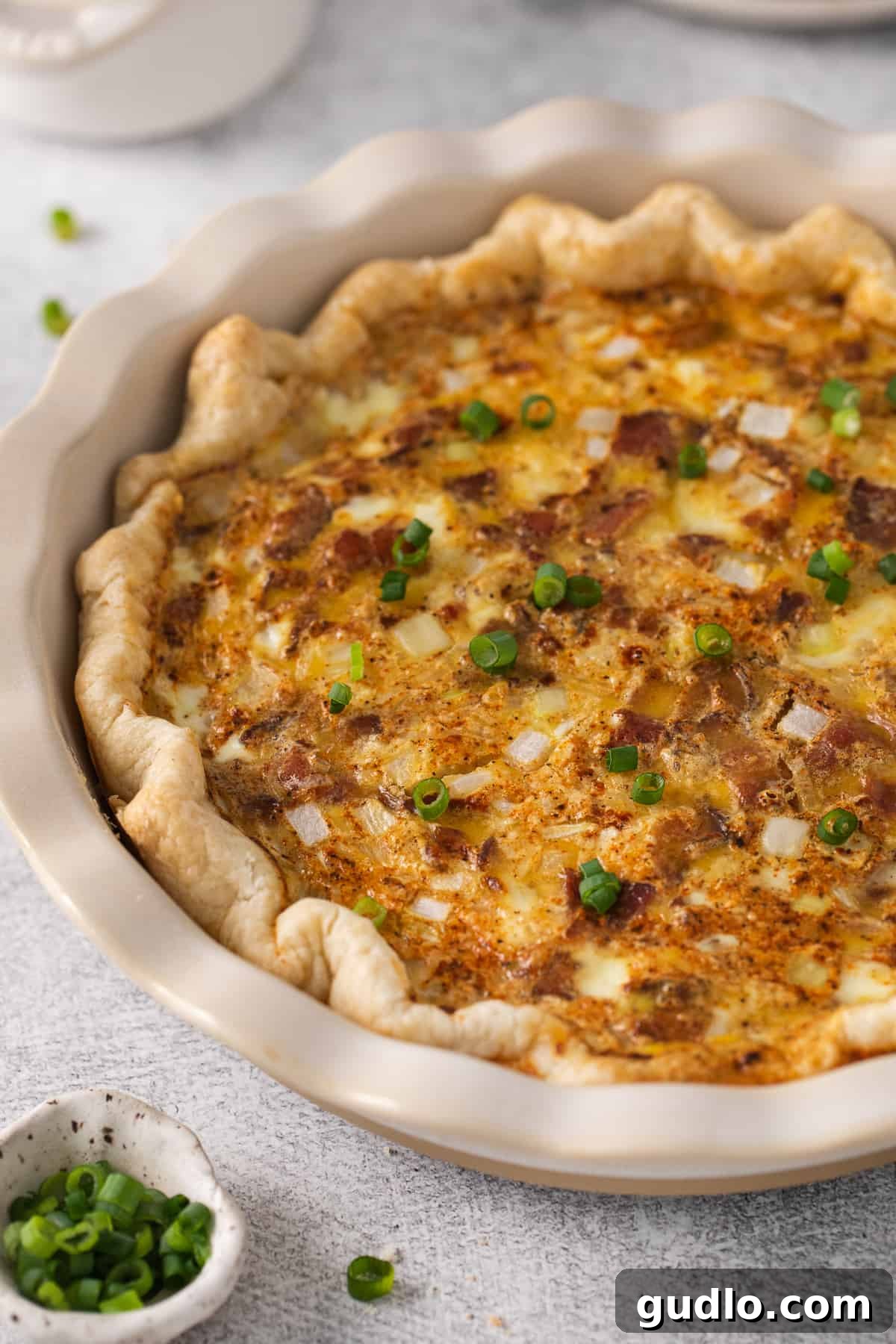 Our easy Quiche Lorraine recipe is the perfect dish for your next brunch! Made with fluffy eggs, cheese, bacon and onions. A freshly baked Quiche Lorraine, garnished with chopped green onions, sitting in its pie plate.
