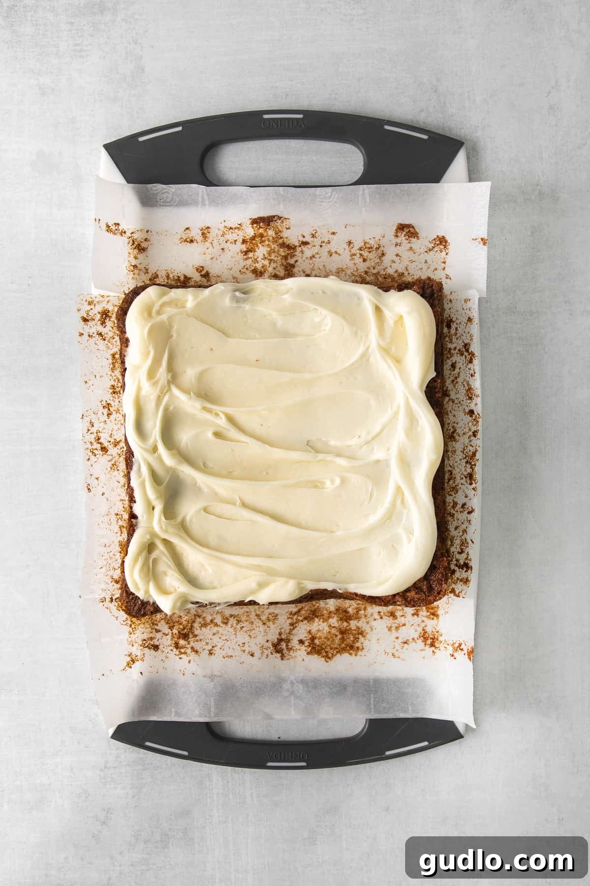 A freshly baked sheet of carrot cake bars covered in cream cheese frosting and decorated with small piped carrots, resting on a wooden counter.