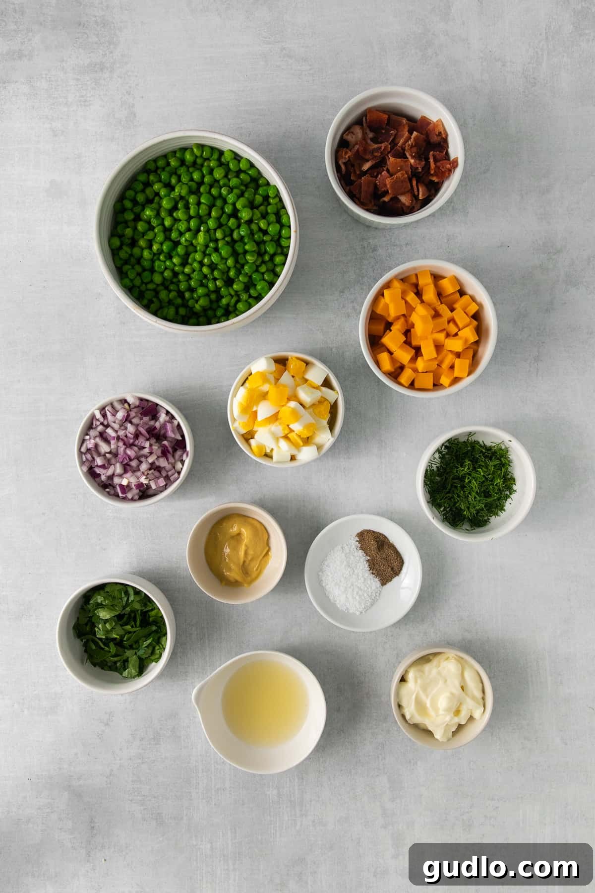 Creamy English Pea Salad 3 Fresh ingredients for English pea salad, including thawed peas, red onion, cheddar cheese, bacon, and hard-boiled eggs, neatly arranged in separate bowls on a kitchen counter.
