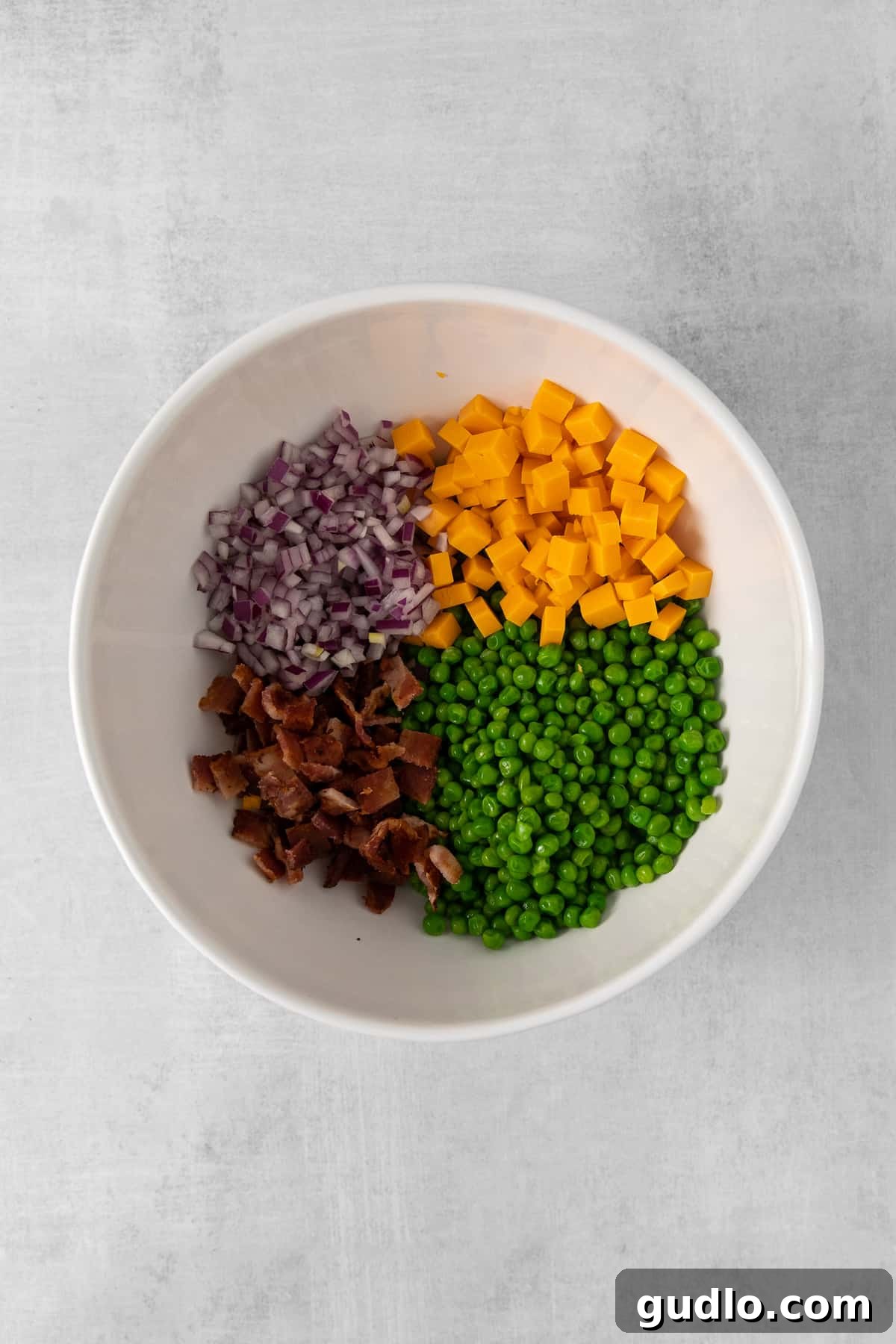 Creamy English Pea Salad 4 A large mixing bowl containing thawed green peas, minced red onion, and diced sharp cheddar cheese, awaiting the creamy dressing and bacon.