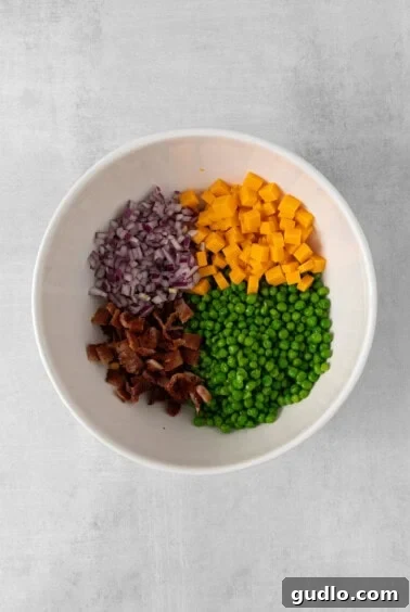 Creamy English Pea Salad 9 Thawed peas, red onion, cheddar cheese, and bacon in a white bowl, ready for mixing.