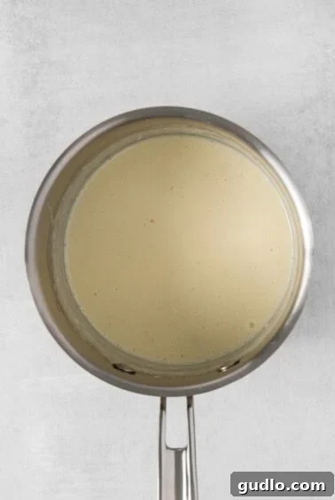 A pan with a white liquid, likely heavy cream and melting cheese, on a white background.