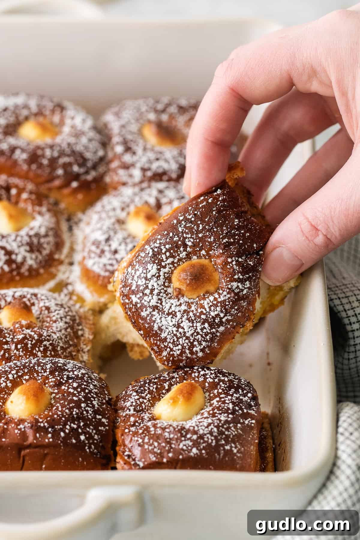 Serving Stuffed Hawaiian Roll French Toast A hand reaching for a piece of Stuffed Hawaiian Roll French Toast from a casserole dish, ready to enjoy.