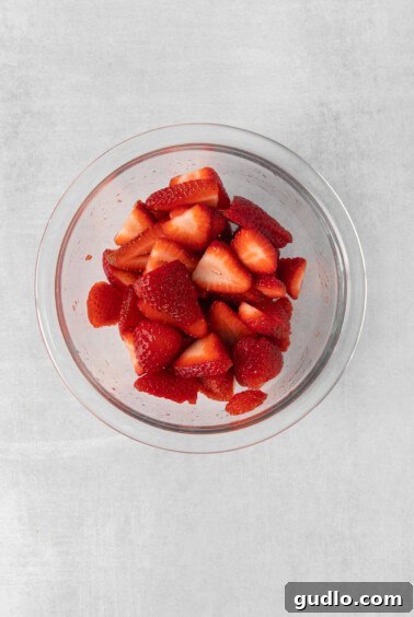 Sliced strawberries in a glass bowl on a grey background, tossed with lemon juice.
