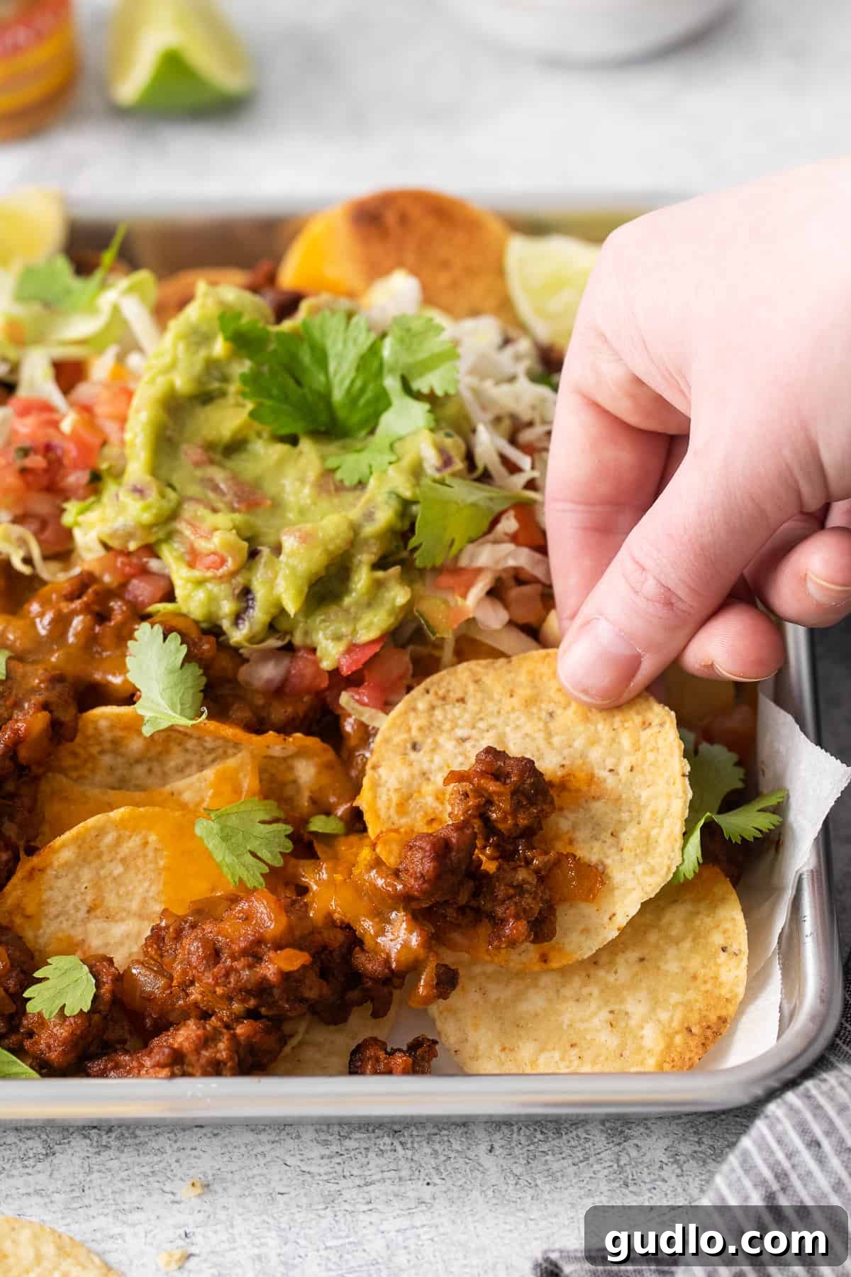 A hand grabbing a tortilla chip with ground beef and melted cheese on it, from a tray of nachos.