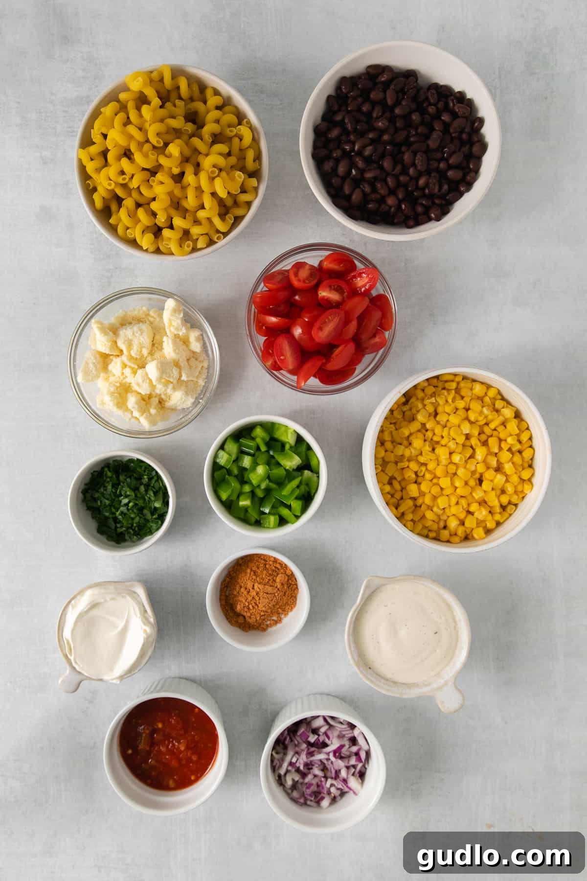 Fiesta Pasta Salad 3 Assortment of fresh ingredients for Mexican pasta salad laid out in individual bowls, including cooked pasta, black beans, corn, bell peppers, cherry tomatoes, and various dressing components.