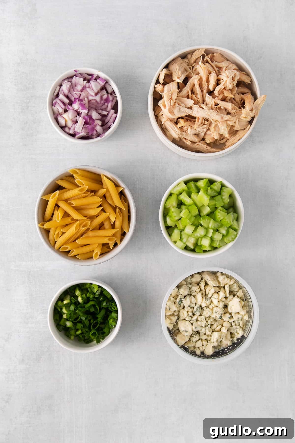 Various fresh ingredients for buffalo chicken pasta salad laid out in individual bowls, including cooked penne pasta, shredded chicken, diced celery, minced red onion, sliced green onions, and blue cheese crumbles.
