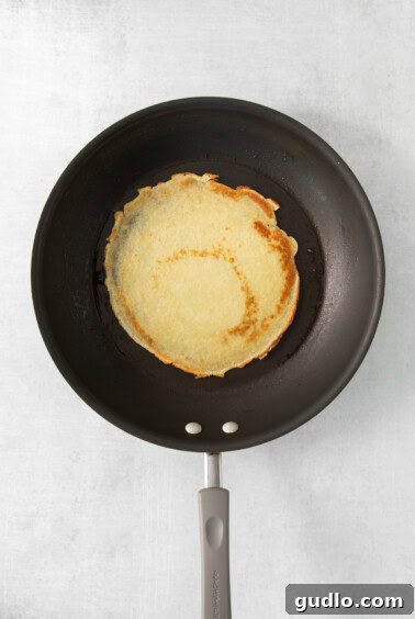 A thin crepe cooking in a nonstick pan, showing its golden-brown edges.