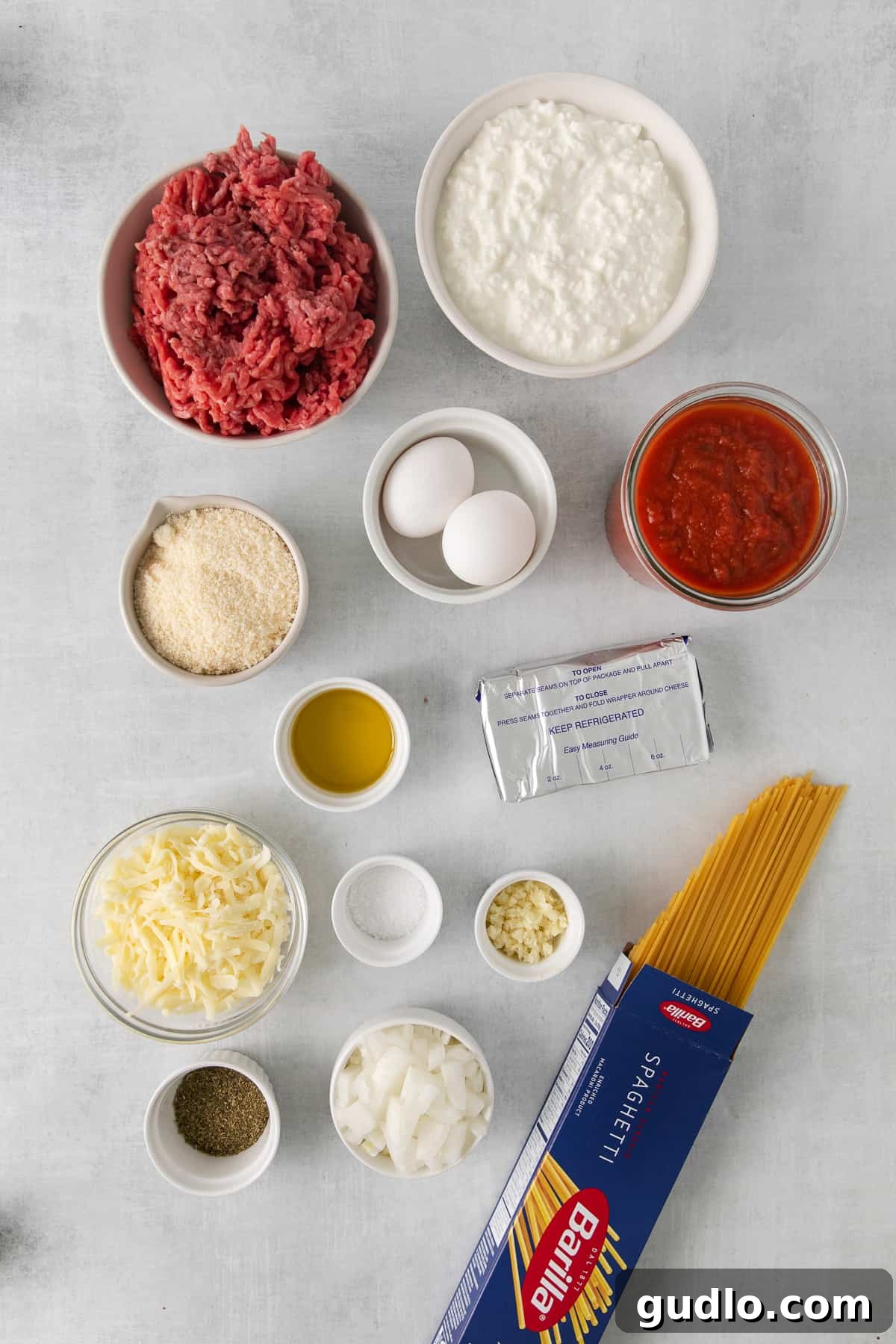 Various fresh ingredients for Million Dollar Spaghetti laid out in small bowls on a counter, including cheeses, spices, onions, and garlic, ready for preparation.