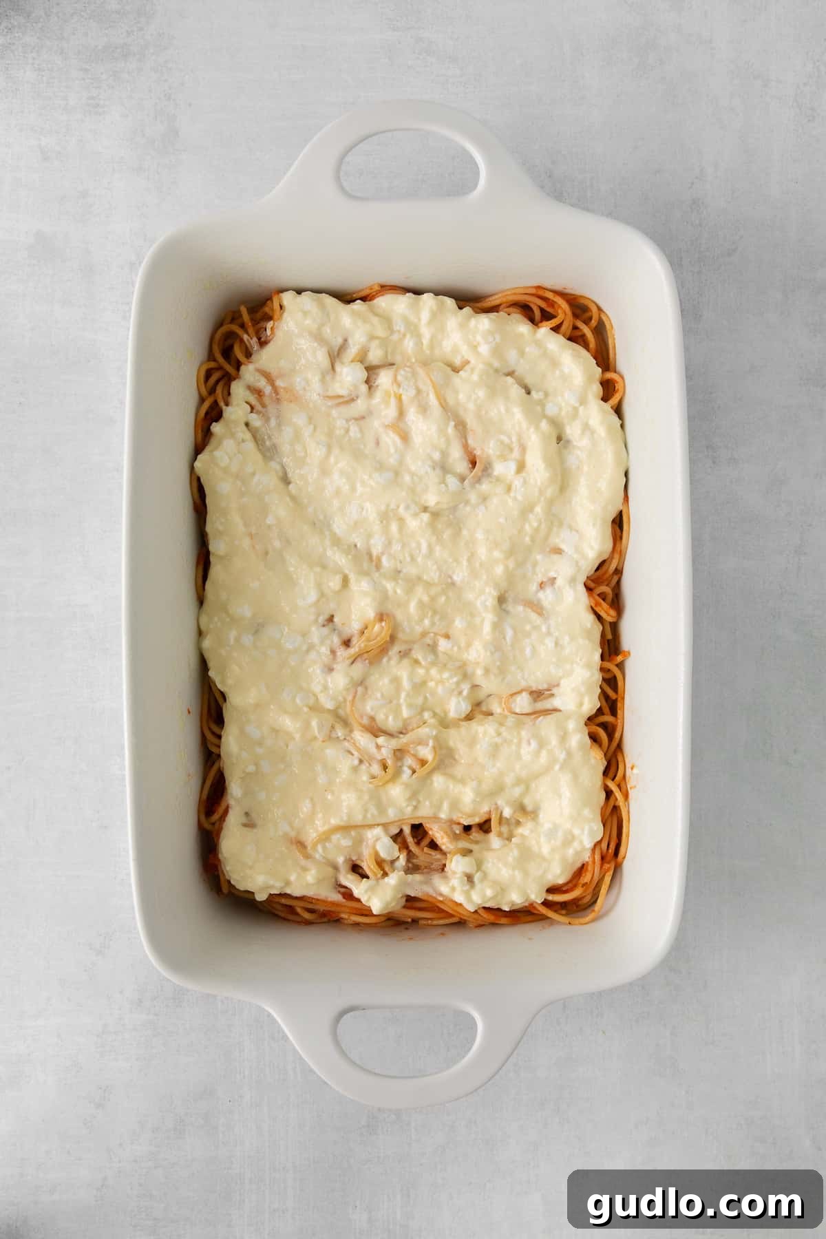 A baking dish filled with spaghetti coated in a creamy white cheese mixture, forming an intermediate layer of the Million Dollar Spaghetti casserole before the final meat and mozzarella topping.