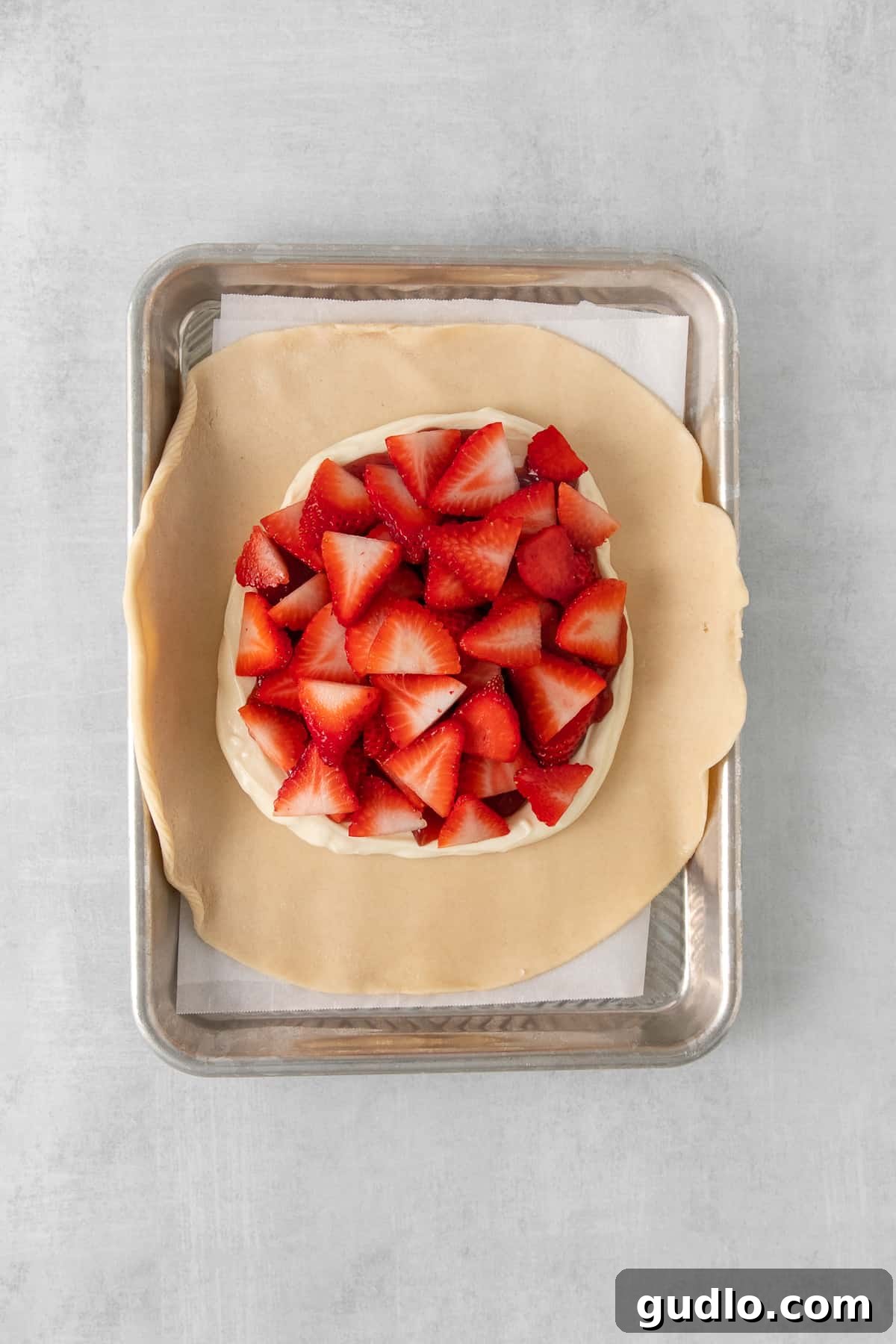 A vibrant display of sliced fresh strawberries meticulously arranged atop a creamy white filling and a layer of strawberry jam, all nestled within a pre-made pie crust on a baking sheet.