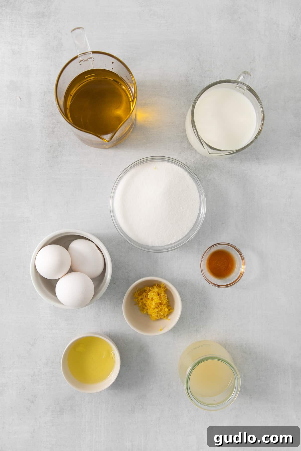 Luminous Lemon Olive Oil Cake 3 A collection of essential ingredients for baking a lemon olive oil cake, neatly arranged in small bowls on a white background, including flour, sugar, lemons, and a bottle of olive oil.
