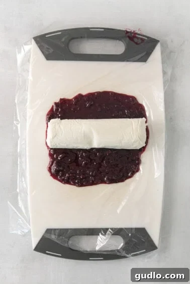 A close-up shot of a goat cheese log being meticulously wrapped in a rectangular layer of rich blueberry compote using plastic wrap on a countertop.