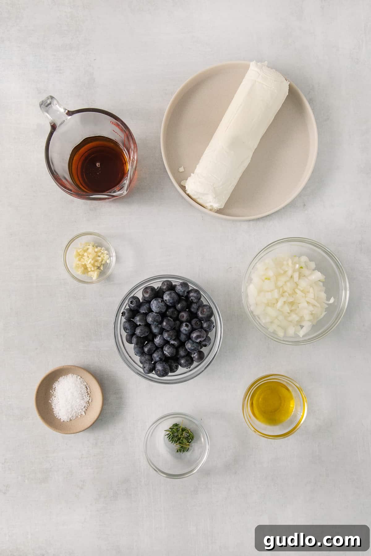 An organized display of all the fresh ingredients required for making the blueberry goat cheese appetizer: a log of goat cheese, fresh blueberries, maple syrup, olive oil, yellow onion, garlic, and thyme sprigs, neatly arranged on a white surface.