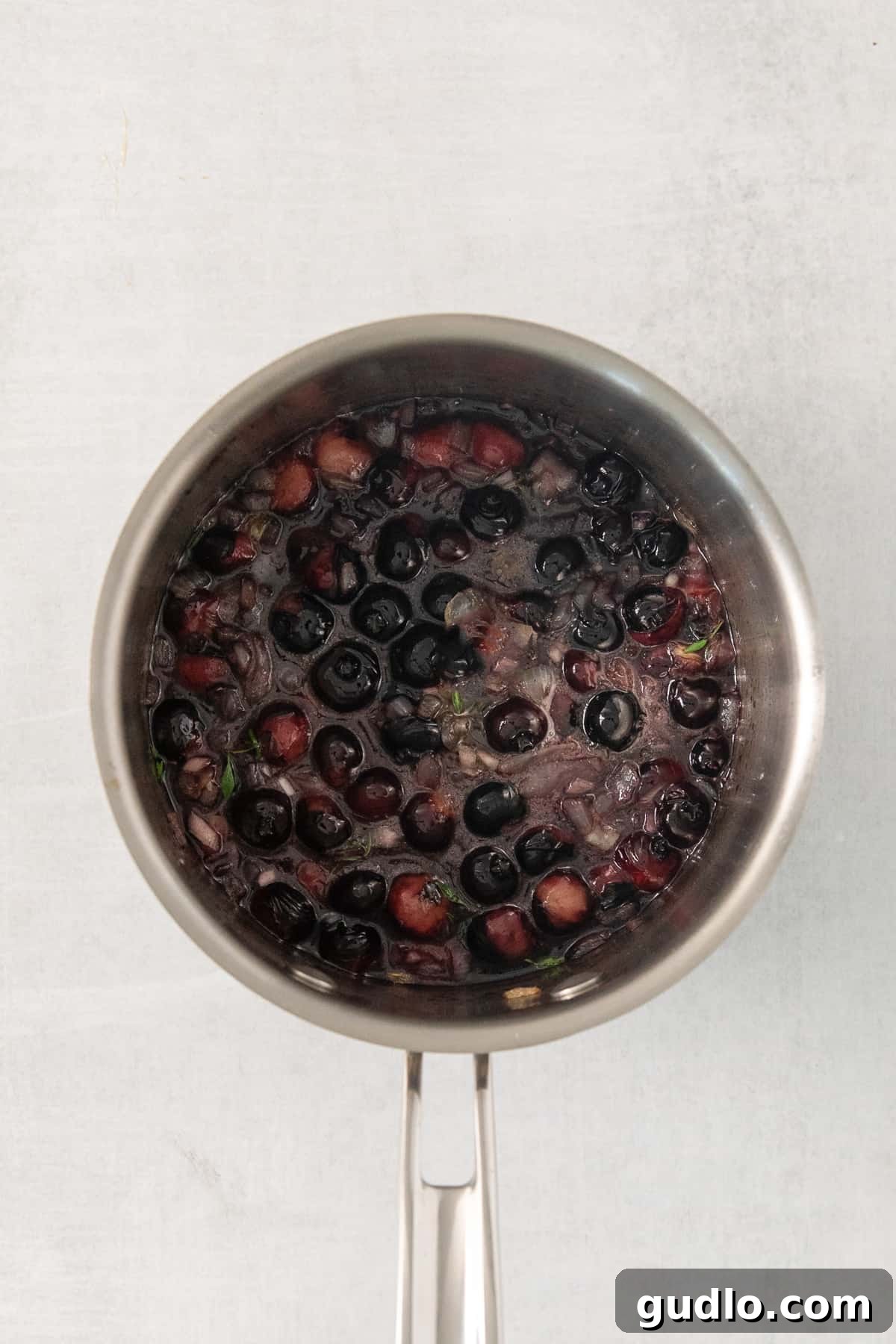 Fresh blueberries simmering gently in a saucepan with minced onion, garlic, maple syrup, and sprigs of thyme, creating a rich and aromatic compote for the goat cheese appetizer.