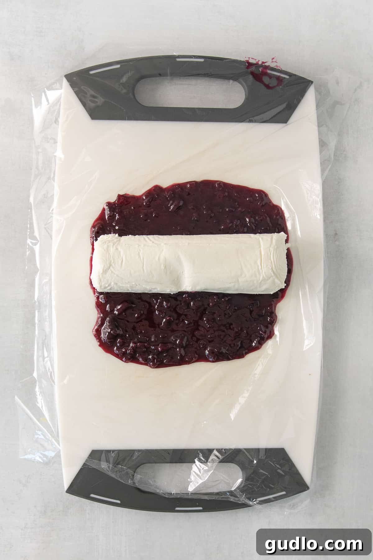 The vibrant blueberry compote cooling in a bowl next to a fresh log of plain goat cheese, illustrating the two main components of the appetizer before assembly.