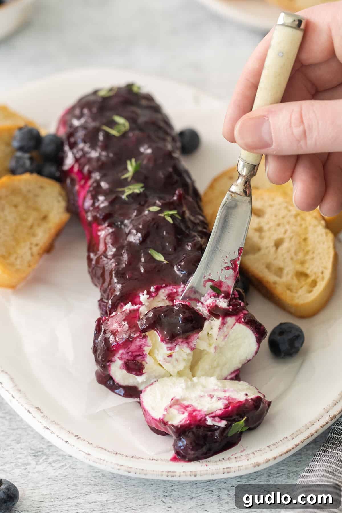 A close-up of a slice of blueberry goat cheese log on a white plate, revealing the creamy goat cheese center and the vibrant purple compote layer, with a knife poised for serving and a sprig of thyme for garnish.