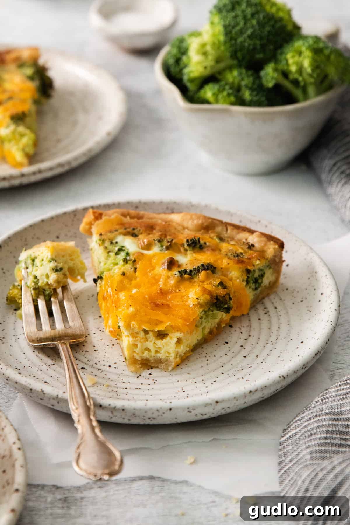 A perfect slice of broccoli cheddar quiche on a white plate, with a fork ready to enjoy.
