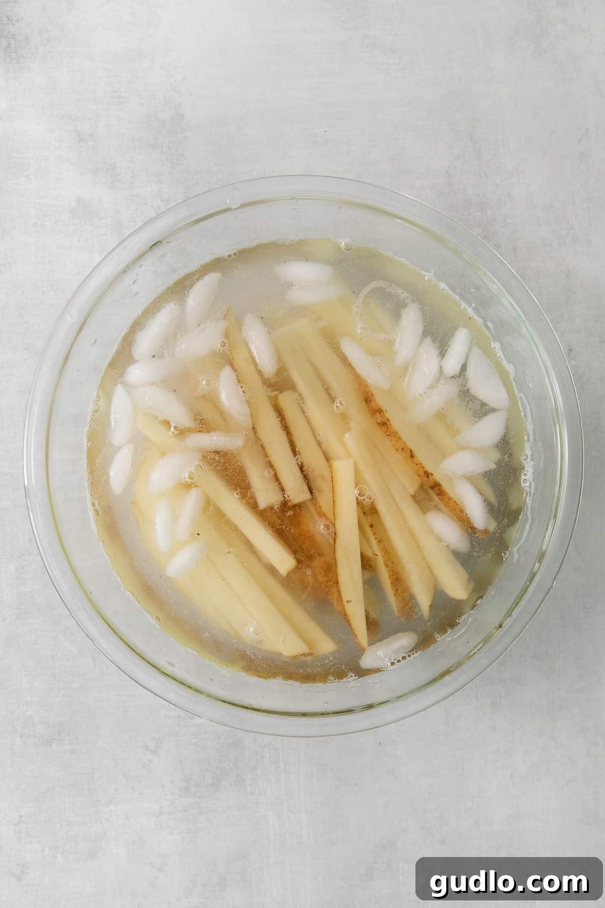 A glass bowl filled with freshly cut russet potato sticks soaking in ice water, a crucial step for achieving extra crispy French fries.