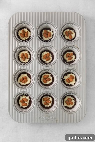 A muffin tin with twelve coconut mounds, each topped with almonds, ready for the final chocolate coating.