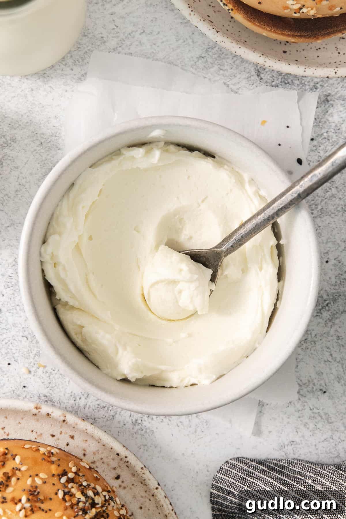 A beautifully textured bowl of homemade cream cheese, ready to be spread, with a spoon and a fresh baguette beside it.