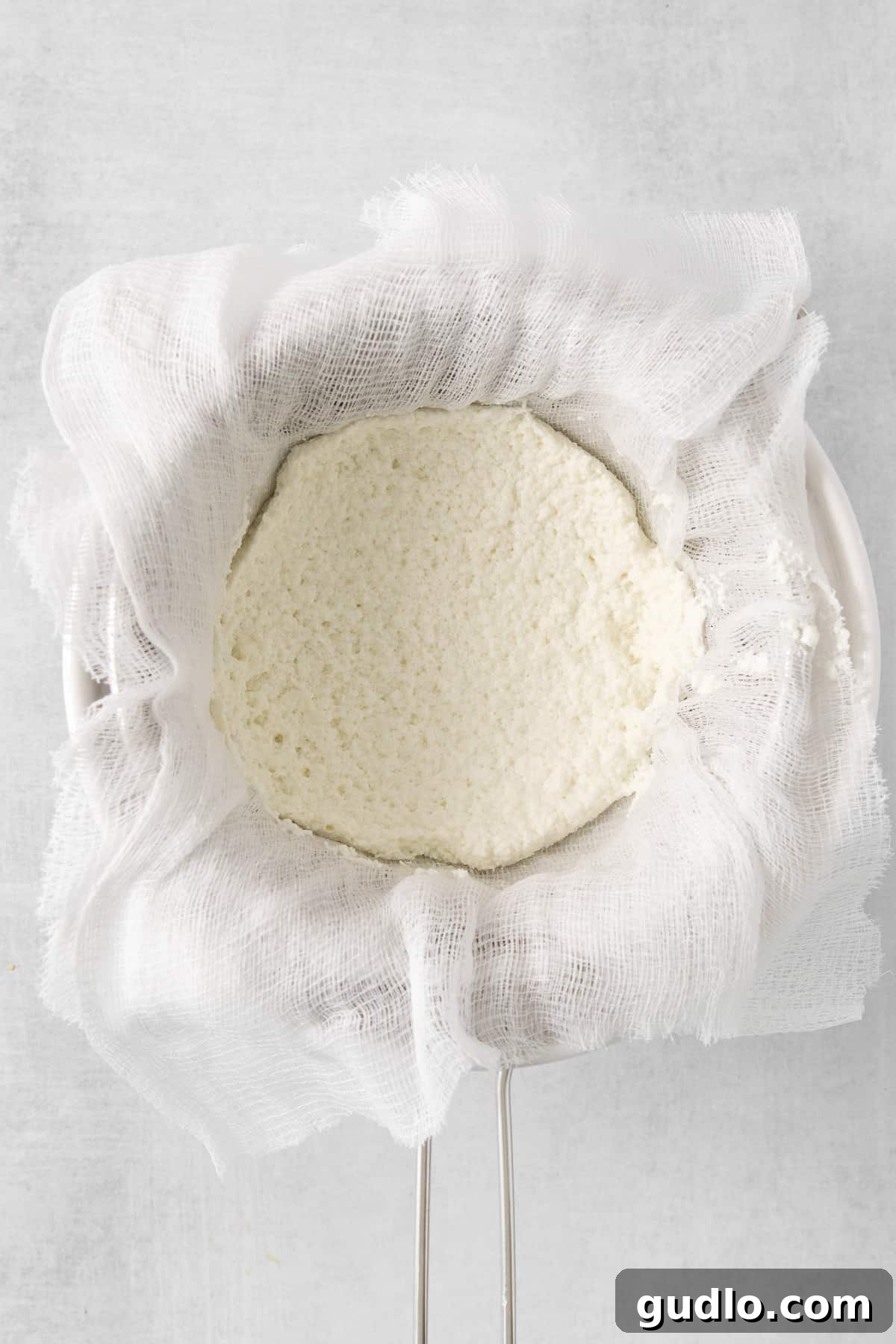 Fresh ricotta cheese delicately held in a cloth bag, dripping excess liquid, on a clean white background, illustrating the straining process.