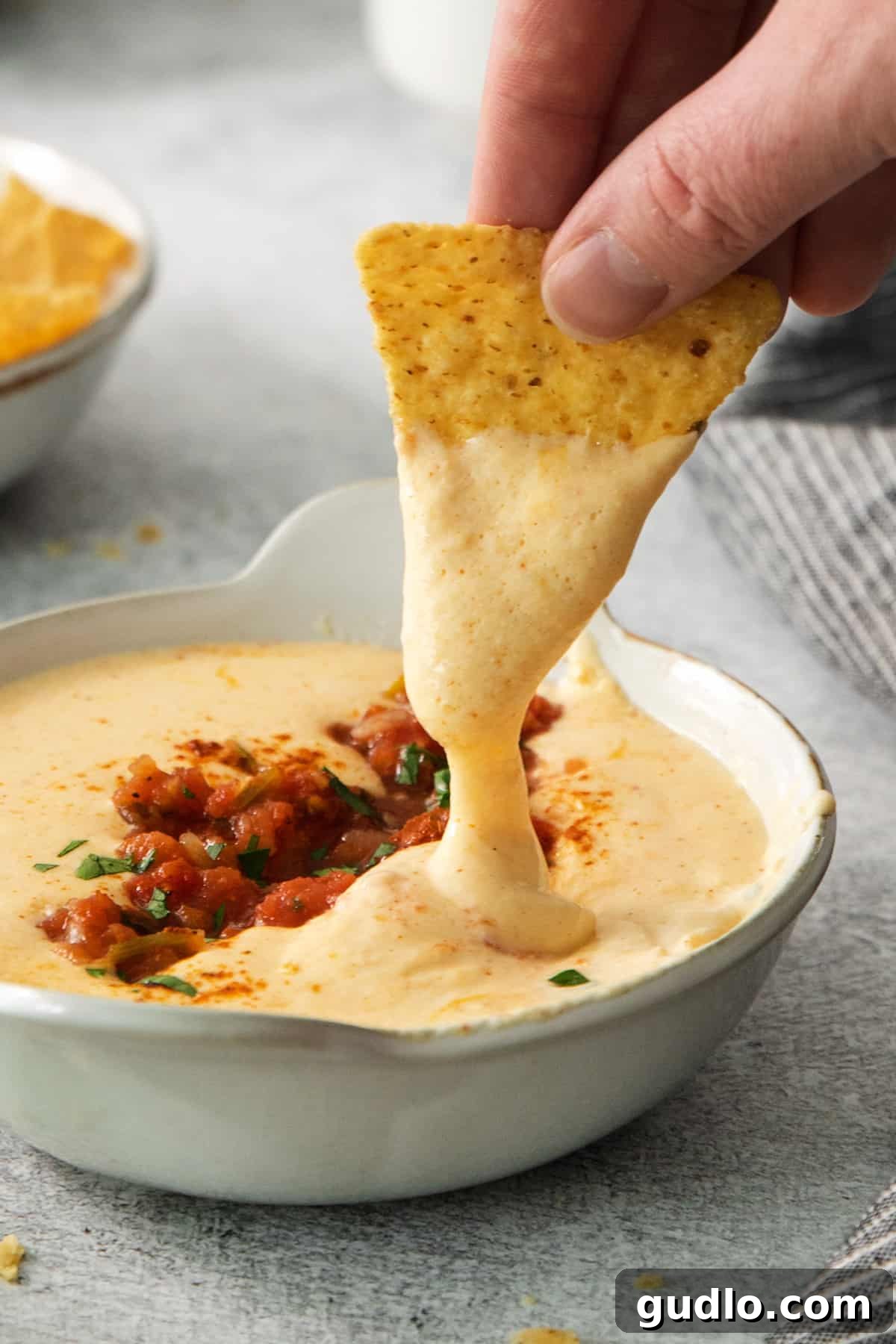 Creamy Protein Cottage Queso 2 A person enthusiastically dipping a golden tortilla chip into a generously filled bowl of creamy, warm cottage cheese queso, showcasing its rich texture.