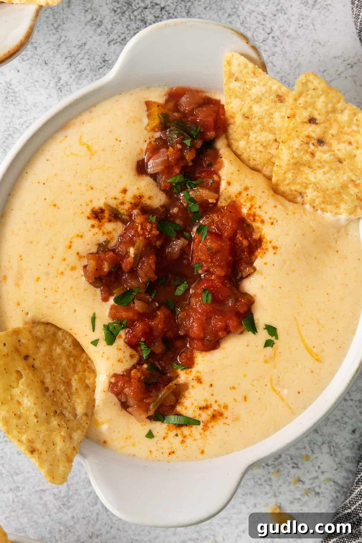 Creamy Protein Cottage Queso 6 A mouthwatering bowl of golden cottage cheese queso, generously garnished with fresh cilantro and a dollop of pico de gallo, surrounded by a scattering of crispy tortilla chips, inviting you to dip in.