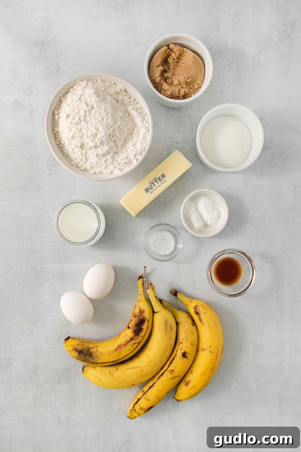 Decadent Cream Cheese Banana Bread Muffins 3 Fresh ingredients for banana bread muffins laid out neatly on a grey background, including flour, butter, sugar, bananas, and eggs.