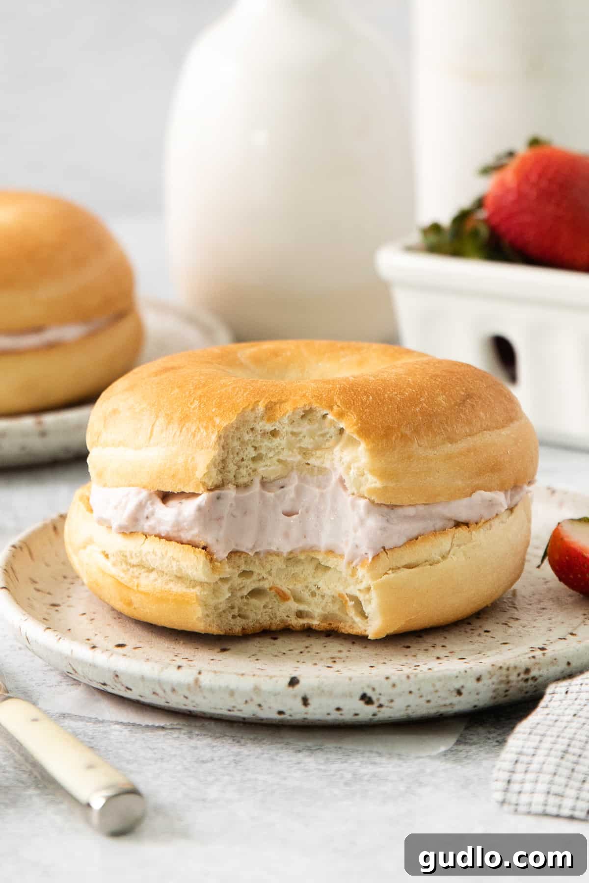 A delectable bagel sandwich, generously spread with creamy strawberry cream cheese and adorned with fresh strawberry slices, presented beautifully on a white plate.