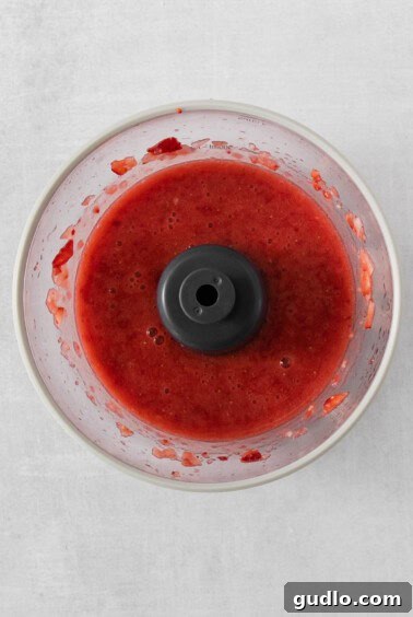 Vibrant red strawberry sauce being processed in a food processor, ready to be incorporated into the creamy cheese base.