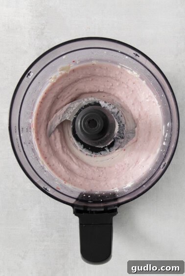 A food processor with a swirling pink liquid, indicating the perfect blend of creamy cheese and strawberry puree.