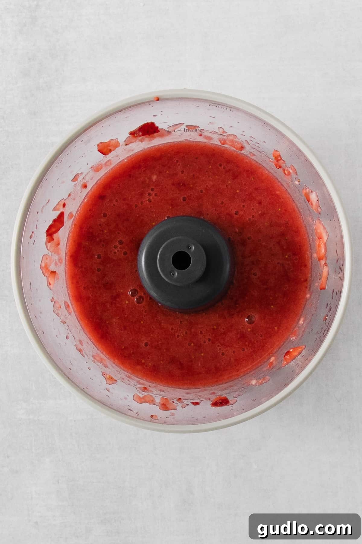 Vibrant red strawberry sauce being processed in a food processor, ready to be incorporated into the creamy cheese base.
